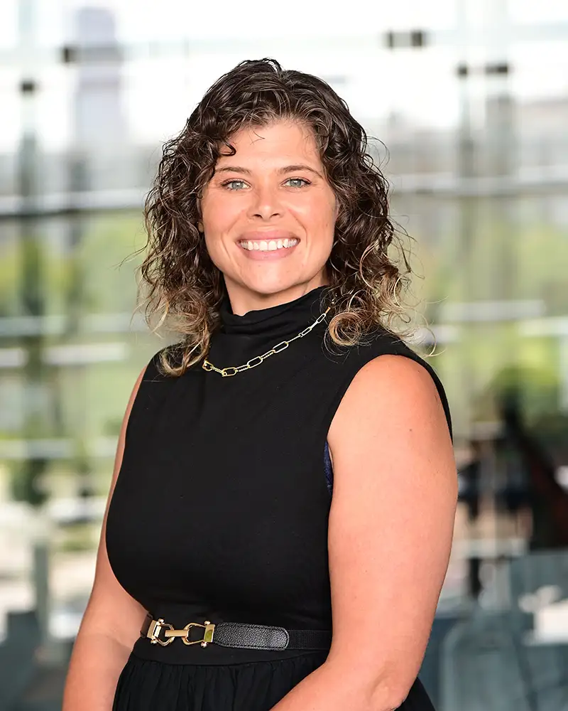 Mary-Kate in black dress and necklace, smiling at camera, blurred cityscape behind