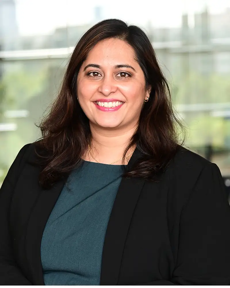 Angira in a black suit jacket and green top smiling at camera, blurred cityscape behind