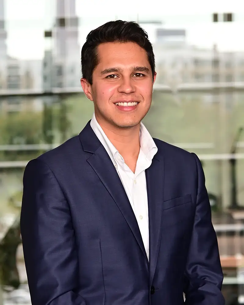 Hans in suit and white collared shirt, no tie, smiling with a blurred cityscape behind him